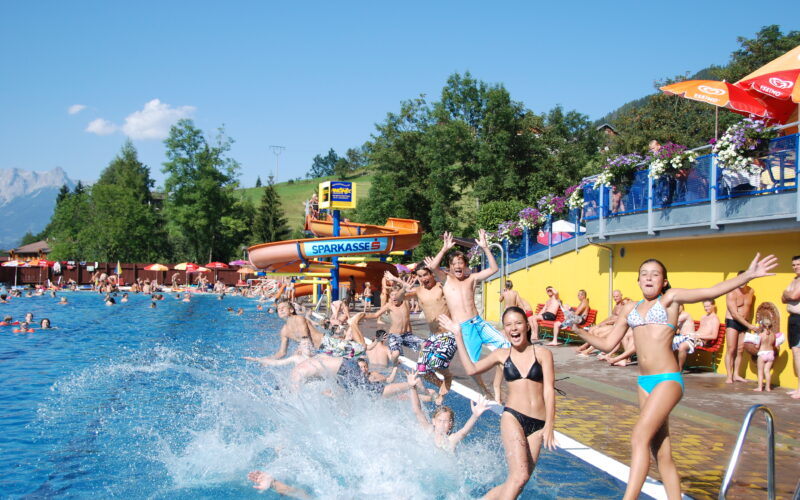 Eine Gruppe von Kindern und Jugendlichen springt an einem sonnigen Tag in ein Freibad, während sich die Leute am Beckenrand entspannen und eine Wasserrutsche im Hintergrund zu sehen ist.