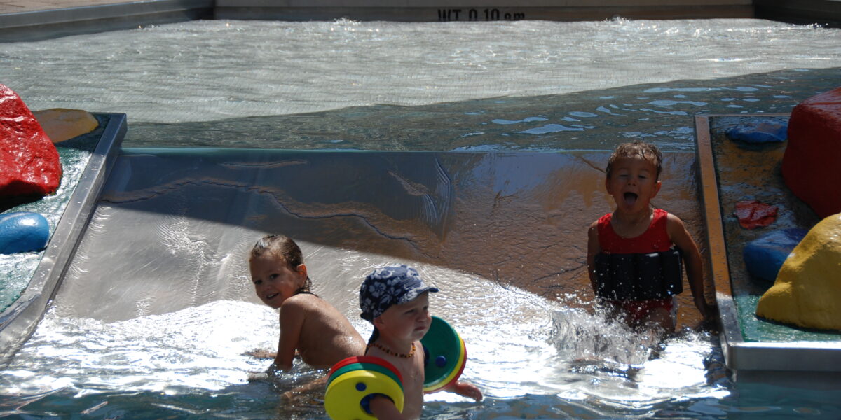 Drei kleine Kinder spielen im seichten Wasser eines Planschbeckens mit kleinen bunten Strukturen und Wasser, das über eine Rampe fließt.