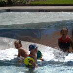 Drei kleine Kinder spielen im seichten Wasser eines Planschbeckens mit kleinen bunten Strukturen und Wasser, das über eine Rampe fließt.