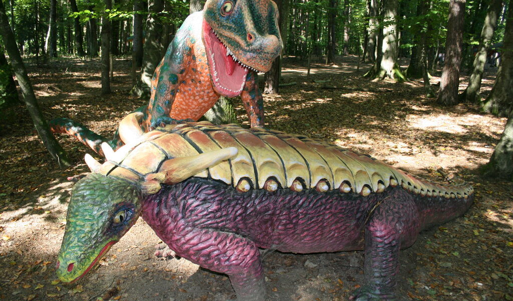 Zwei farbenfrohe Dinosaurierstatuen, eine über der anderen stehend, werden in einem bewaldeten Außenbereich ausgestellt, durch dessen Bäume das Sonnenlicht fällt.