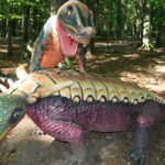 Zwei farbenfrohe Dinosaurierstatuen, eine über der anderen stehend, werden in einem bewaldeten Außenbereich ausgestellt, durch dessen Bäume das Sonnenlicht fällt.