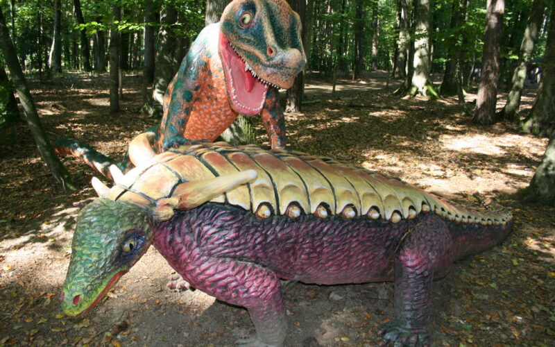 Zwei farbenfrohe Dinosaurierstatuen, eine über der anderen stehend, werden in einem bewaldeten Außenbereich ausgestellt, durch dessen Bäume das Sonnenlicht fällt.