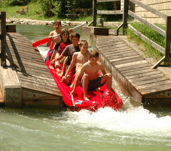 An einem sonnigen Tag fahren fünf Kinder auf einem roten Schlauchboot eine Wasserrutsche hinunter und planschen in einem Becken am Boden.