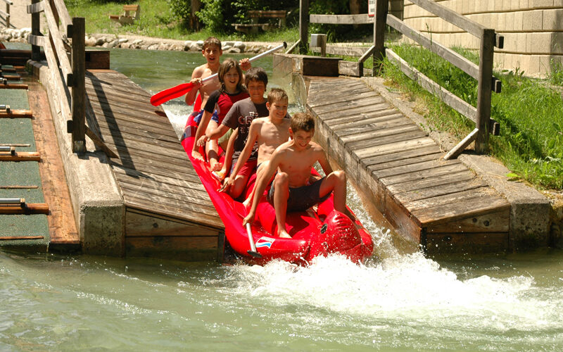 An einem sonnigen Tag fahren fünf Kinder auf einem roten Schlauchboot eine Wasserrutsche hinunter und planschen in einem Becken am Boden.