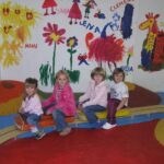 Vier kleine Kinder sitzen auf einer Holzwippe in einem farbenfrohen Spielzimmer mit Wandgemälden mit Blumen, Tieren und Kindernamen an der Wand.