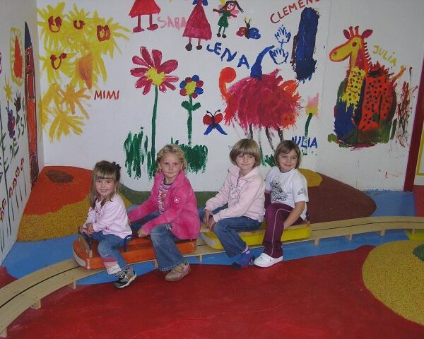 Vier kleine Kinder sitzen auf einer Holzwippe in einem farbenfrohen Spielzimmer mit Wandgemälden mit Blumen, Tieren und Kindernamen an der Wand.