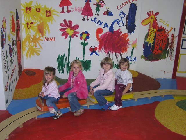 Vier kleine Kinder sitzen auf einer Holzwippe in einem farbenfrohen Spielzimmer mit Wandgemälden mit Blumen, Tieren und Kindernamen an der Wand.