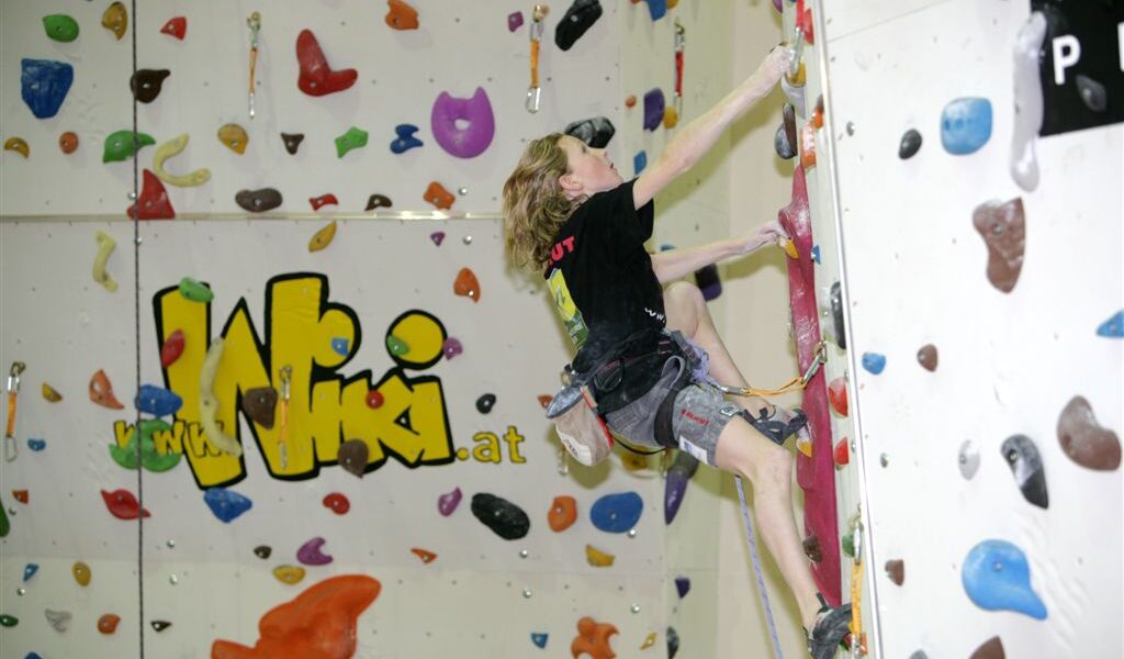 Eine Person in Kletterausrüstung besteigt eine Indoor-Kletterwand mit bunten Griffen; im Hintergrund ist ein großes "WIKI"-Logo zu sehen.