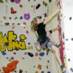 Eine Person in Kletterausrüstung besteigt eine Indoor-Kletterwand mit bunten Griffen; im Hintergrund ist ein großes "WIKI"-Logo zu sehen.