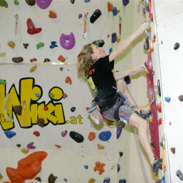 Eine Person in Kletterausrüstung besteigt eine Indoor-Kletterwand mit bunten Griffen; im Hintergrund ist ein großes "WIKI"-Logo zu sehen.