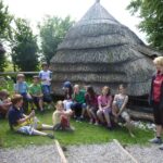 Eine Gruppe von Kindern und ein Erwachsener sitzen und stehen in der Nähe einer strohgedeckten Hütte auf einer Wiese, mit Bäumen im Hintergrund.
