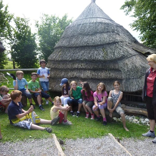Eine Gruppe von Kindern und ein Erwachsener sitzen und stehen in der Nähe einer strohgedeckten Hütte auf einer Wiese, mit Bäumen im Hintergrund.