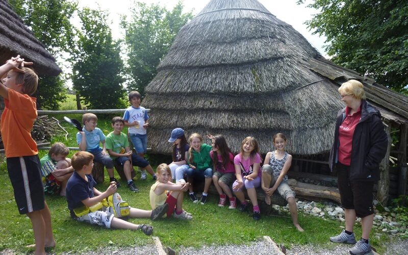 Eine Gruppe von Kindern und ein Erwachsener sitzen und stehen in der Nähe einer strohgedeckten Hütte auf einer Wiese, mit Bäumen im Hintergrund.