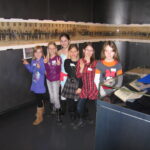 Sechs Kinder stehen in einem Raum zusammen und machen Friedenszeichen vor einer Vitrine und einem langen historischen Foto, das an der Wand hinter ihnen angebracht ist.