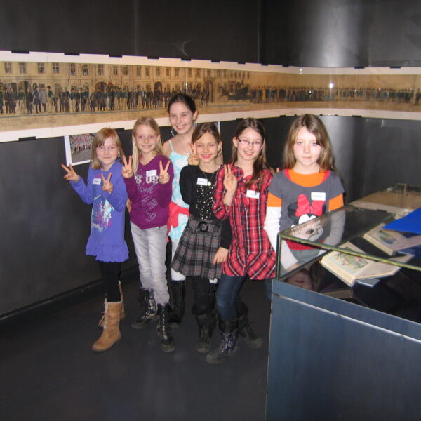 Sechs Kinder stehen in einem Raum zusammen und machen Friedenszeichen vor einer Vitrine und einem langen historischen Foto, das an der Wand hinter ihnen angebracht ist.