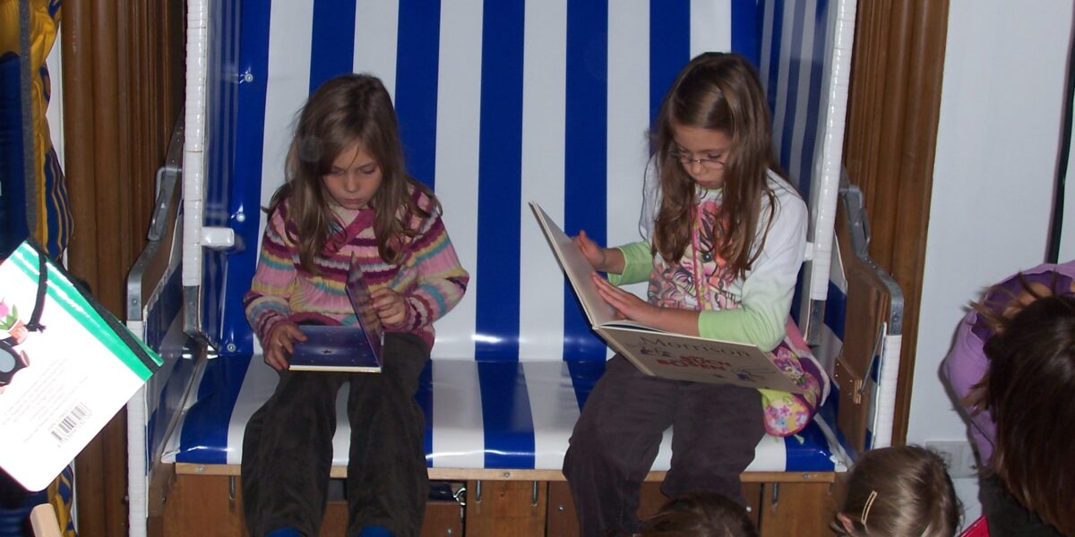Zwei junge Mädchen sitzen auf einer blau-weiß gestreiften Bank und lesen Bücher, während andere Kinder vor ihnen auf dem Boden sitzen oder knien.