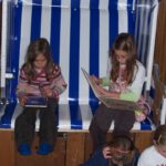 Zwei junge Mädchen sitzen auf einer blau-weiß gestreiften Bank und lesen Bücher, während andere Kinder vor ihnen auf dem Boden sitzen oder knien.
