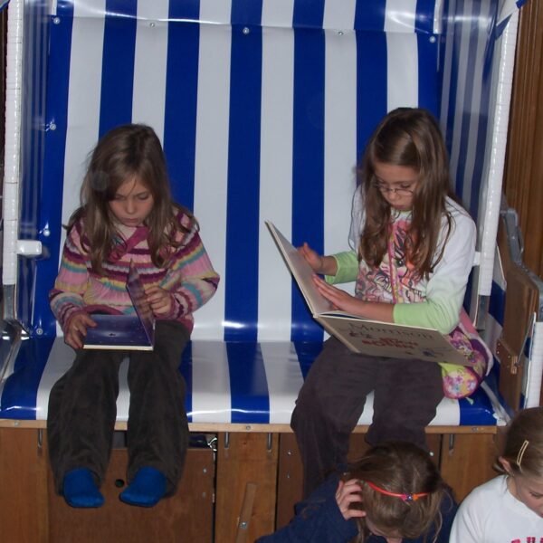 Zwei junge Mädchen sitzen auf einer blau-weiß gestreiften Bank und lesen Bücher, während andere Kinder vor ihnen auf dem Boden sitzen oder knien.