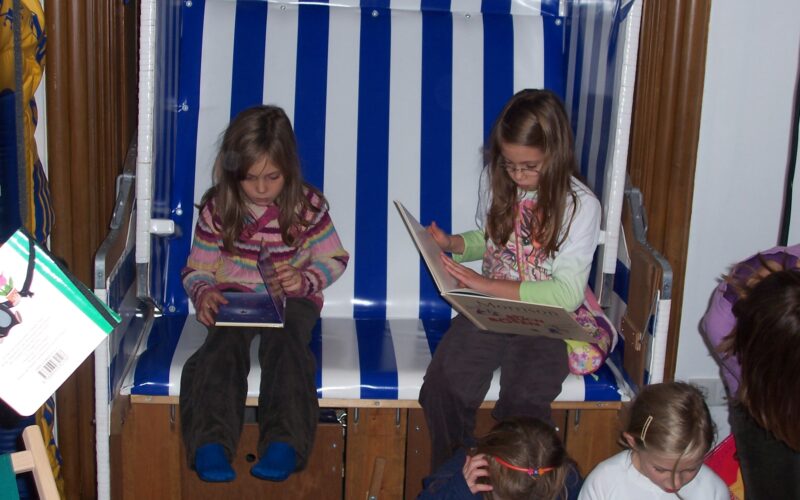 Zwei junge Mädchen sitzen auf einer blau-weiß gestreiften Bank und lesen Bücher, während andere Kinder vor ihnen auf dem Boden sitzen oder knien.