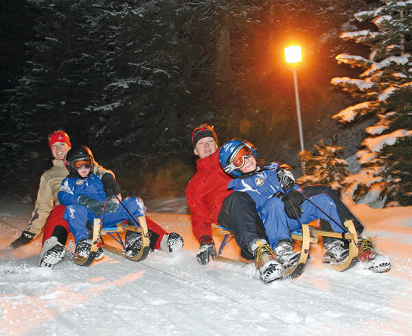 Zwei Erwachsene und zwei Kinder in Winterkleidung fahren nachts auf einem verschneiten Weg Schlitten, mit Bäumen und einer Straßenlaterne im Hintergrund.
