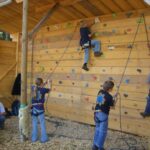 Kinder mit Klettergurten klettern und bereiten sich darauf vor, eine hölzerne Indoor-Kletterwand mit bunten Griffen zu erklimmen; Erwachsene beaufsichtigen sie am Rand.