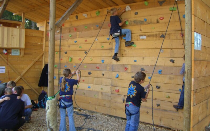 Kinder mit Klettergurten klettern und bereiten sich darauf vor, eine hölzerne Indoor-Kletterwand mit bunten Griffen zu erklimmen; Erwachsene beaufsichtigen sie am Rand.