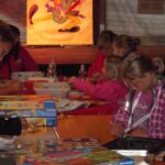 Kinder und Erwachsene sitzen an einem Tisch, spielen Brettspiele und arbeiten an Aktivitäten, wobei Spielkisten und Zubehör in einem belebten Raum verteilt sind.