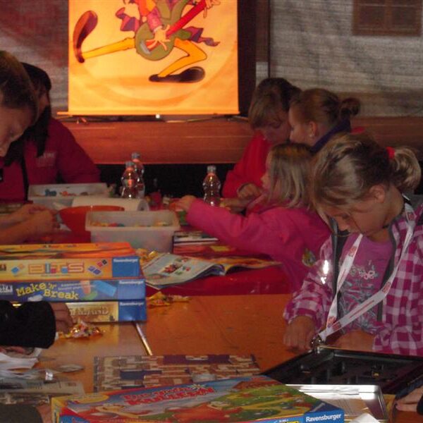 Kinder und Erwachsene sitzen an einem Tisch, spielen Brettspiele und arbeiten an Aktivitäten, wobei Spielkisten und Zubehör in einem belebten Raum verteilt sind.