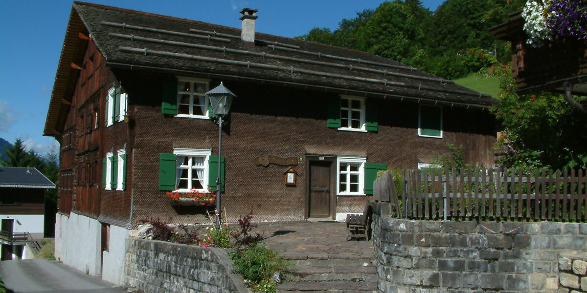Ein traditionelles Holzhaus mit grünen Fensterläden und Blumenkästen, Steintreppen und einer Steinmauer vor dem Hintergrund von Bäumen und einem blauen Himmel.