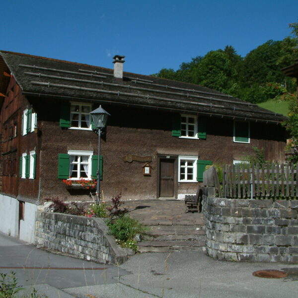 Ein traditionelles Holzhaus mit grünen Fensterläden und Blumenkästen, Steintreppen und einer Steinmauer vor dem Hintergrund von Bäumen und einem blauen Himmel.
