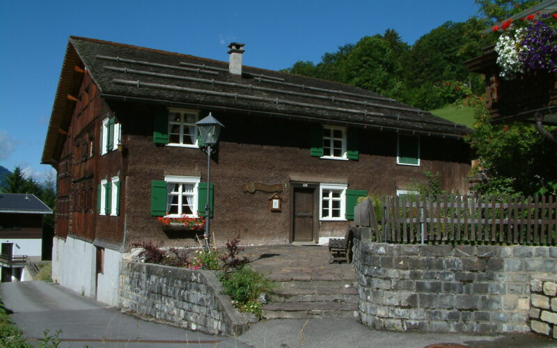 Ein traditionelles Holzhaus mit grünen Fensterläden und Blumenkästen, Steintreppen und einer Steinmauer vor dem Hintergrund von Bäumen und einem blauen Himmel.