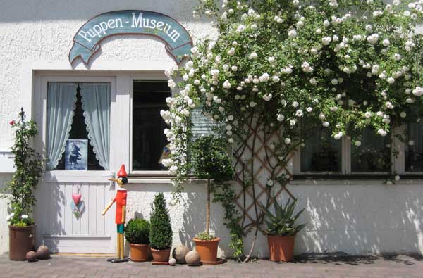 Der Eingang zu einem Gebäude mit einem Schild "Puppen-Museum", Topfpflanzen, einer hölzernen Pinocchio-Figur und weißen Blumen an einem Spalier neben der Tür.
