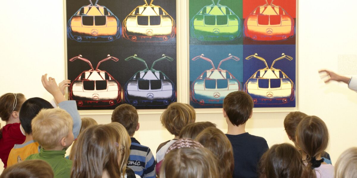 Eine Gruppe von Kindern steht vor zwei bunten Pop-Art-Gemälden von Autos, die nebeneinander in einer Galerie ausgestellt sind.