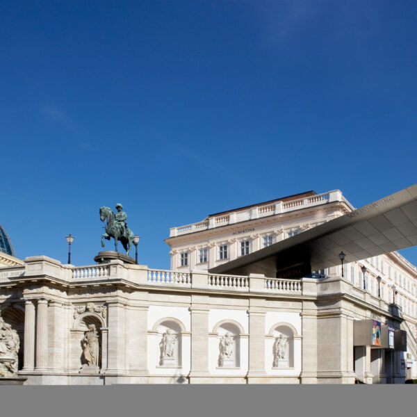 Ein großes Reiterstandbild steht auf einem klassischen Steinbau mit Bögen und Skulpturen neben einem modernen, kantigen Dach unter einem klaren blauen Himmel.