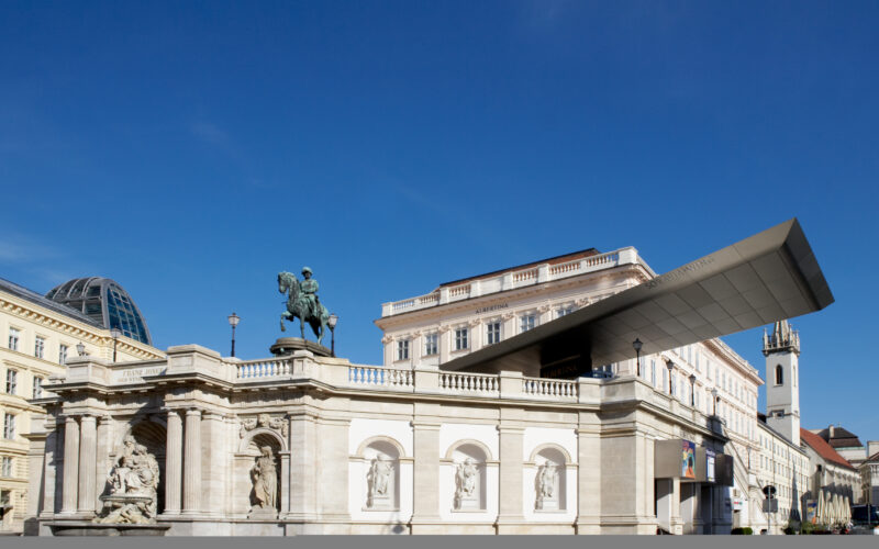 Ein modernes Metalldach erstreckt sich über einer historischen Reiterstatue und einem Steindenkmal mit klassischen Skulpturen, die vor der Kulisse städtischer Gebäude unter einem klaren blauen Himmel stehen.