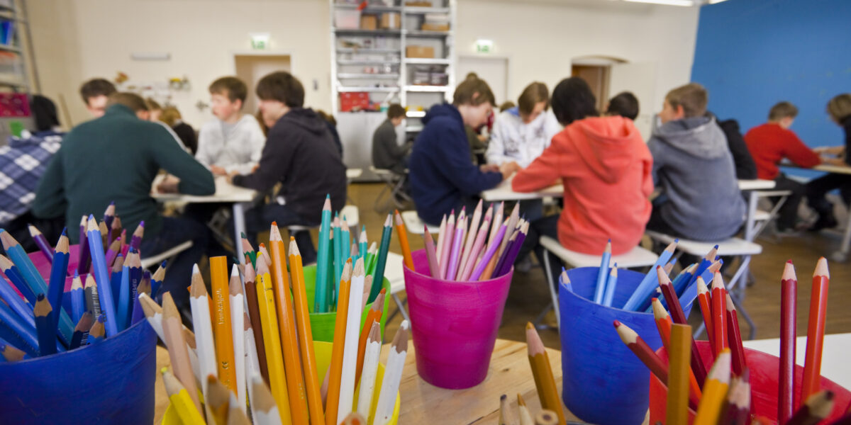 Buntstifte in Bechern auf einem Tisch im Vordergrund, mit Gruppen von Schülern, die in einem Klassenzimmer im Hintergrund zusammensitzen und arbeiten.