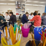 Buntstifte in Bechern auf einem Tisch im Vordergrund, mit Gruppen von Schülern, die in einem Klassenzimmer im Hintergrund zusammensitzen und arbeiten.