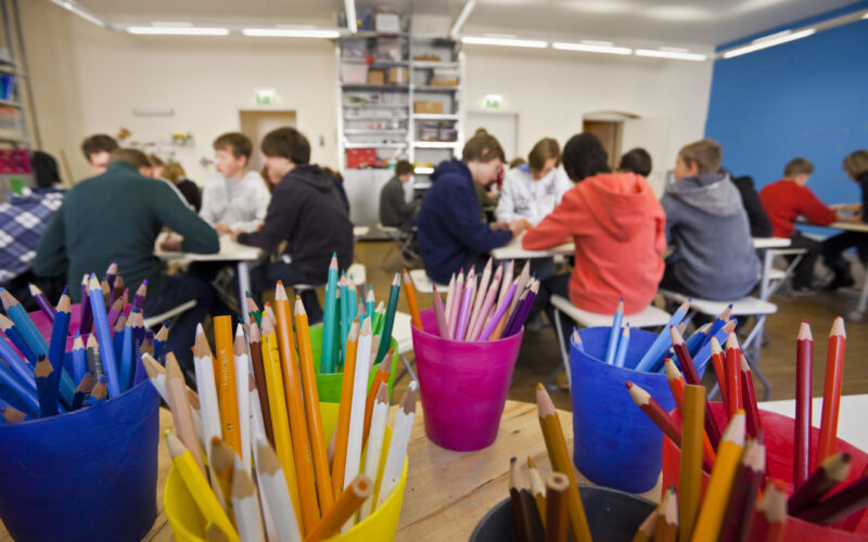 Buntstifte in Bechern auf einem Tisch im Vordergrund, mit Gruppen von Schülern, die in einem Klassenzimmer im Hintergrund zusammensitzen und arbeiten.