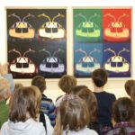 Eine Gruppe von Kindern steht vor zwei bunten Pop-Art-Gemälden von Autos, die nebeneinander an einer Wand hängen.