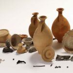 Eine Sammlung von antiken Keramikgefäßen, Schalen, Deckeln, Münzen, Metallgegenständen und Keramikfragmenten, die auf einem schlichten weißen Hintergrund angeordnet sind.