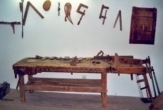 Eine hölzerne Werkbank mit verschiedenen Handwerkzeugen darauf und darüber an der Wand hängenden Tischlerwerkzeugen.