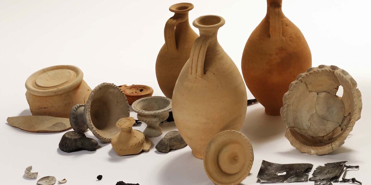 Eine Sammlung von antiken Keramikgefäßen, Keramikscherben, Münzen, Metallfragmenten und kleinen Artefakten auf weißem Hintergrund.