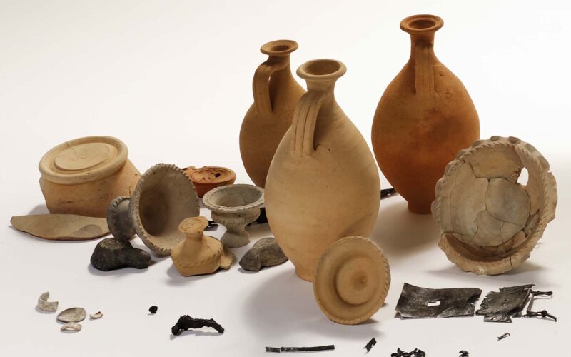 Eine Sammlung von antiken Keramikgefäßen, Keramikscherben, Münzen, Metallfragmenten und kleinen Artefakten auf weißem Hintergrund.