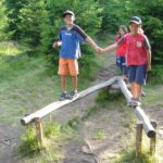 Zwei Jungen stehen händchenhaltend auf einem V-förmigen hölzernen Schwebebalken im Freien, während ein Mädchen hinter ihnen in der Nähe eines Waldes auf dem Boden steht.