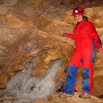 Eine Person in einem rot-blauen Höhlenanzug und einem Helm steht in einer Felsenhöhle neben einem kleinen, fließenden Bach.