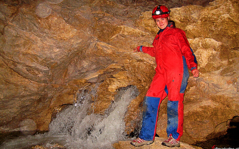 Eine Person in einem rot-blauen Höhlenanzug und einem Helm steht in einer Felsenhöhle neben einem kleinen, fließenden Bach.