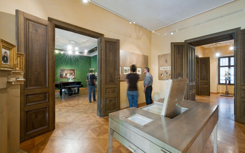 Menschen, die Kunstwerke in einem geräumigen, gut beleuchteten Museumsraum mit Holzböden, großen Türen und Kunstwerken an den Wänden und in einer Vitrine betrachten.