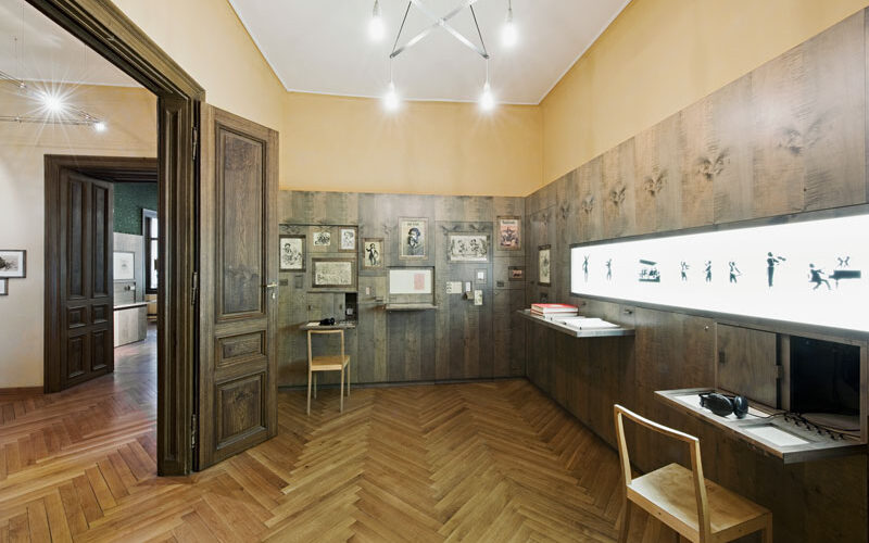 Ein Museumsraum mit Holzböden und -wänden, Vitrinen mit kleinen Skulpturen, gerahmten Kunstwerken, zwei Holzstühlen und Deckenbeleuchtung.