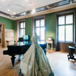 Eleganter historischer Raum mit grünen Tapeten, Holzböden, einem Flügel, einer Orgel und einem ausgestellten historischen Kleid. Große Fenster lassen viel natürliches Licht herein.