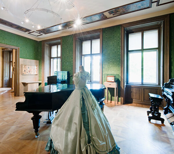 Eleganter historischer Raum mit grünen Tapeten, Holzböden, einem Flügel, einer Orgel und einem ausgestellten historischen Kleid. Große Fenster lassen viel natürliches Licht herein.